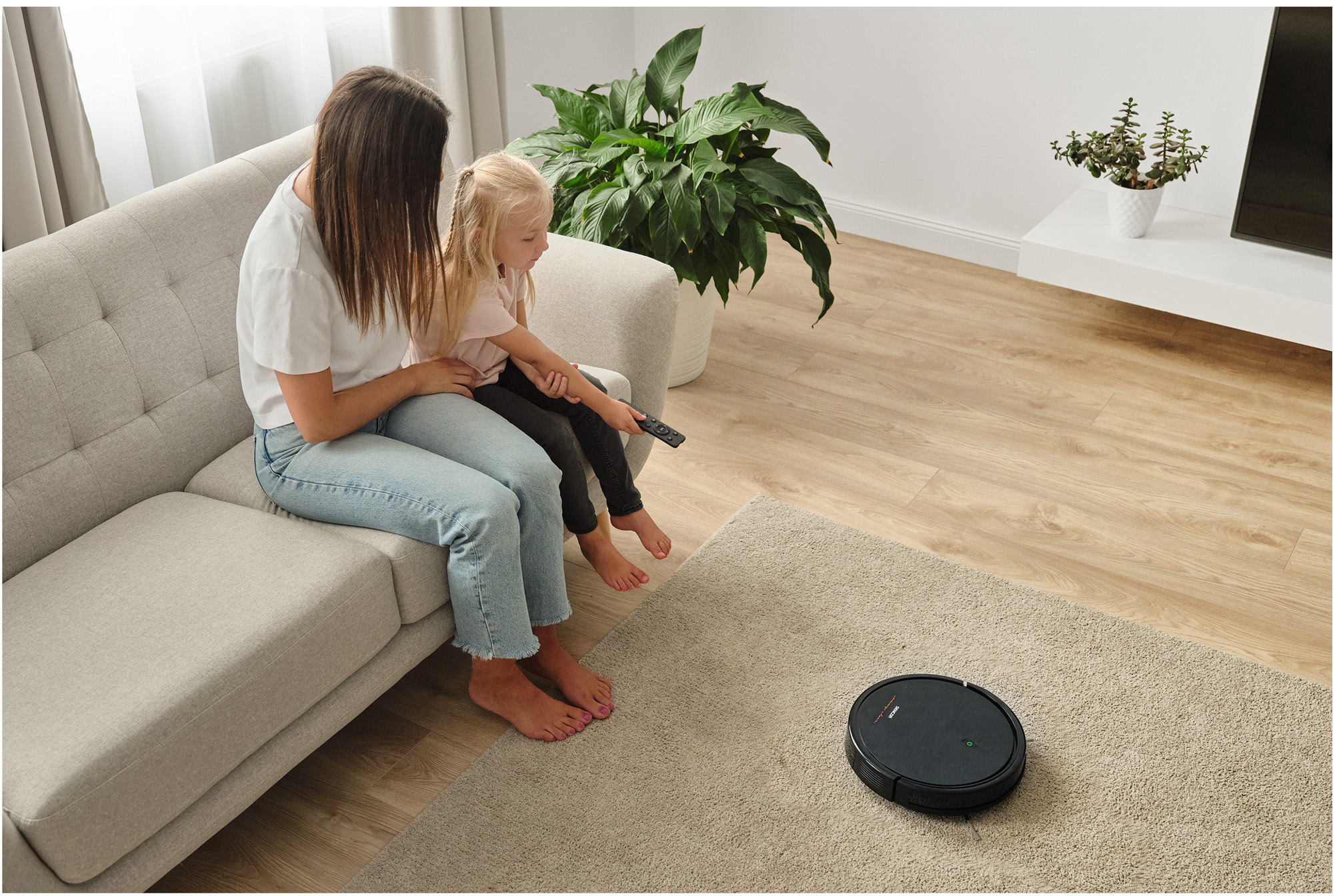 Nő és lánya a kanapén, egy robotporszívót néznek a szőnyegen.