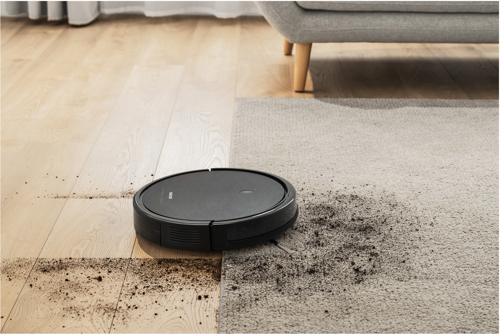 Robotporszívó takarít egy padlót. A fele tiszta, a másik fele törmelékes. Kanapé és szőnyeg a háttérben.