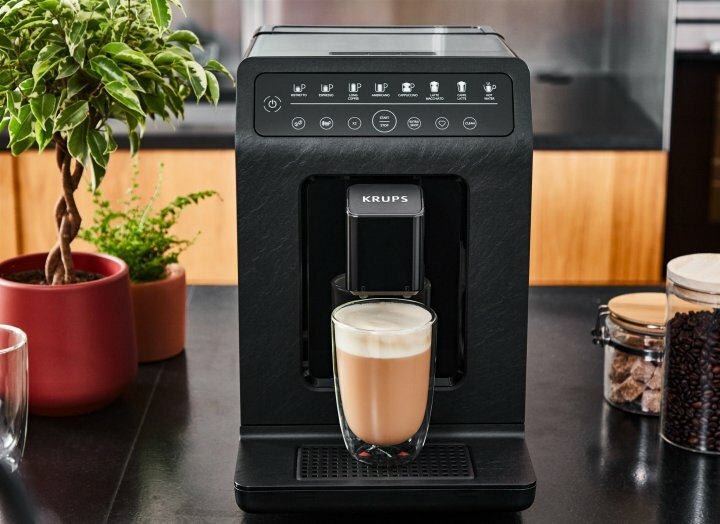 Czarny ekspres do kawy Krups ze szklanką kawy. Obecna jest również roślina i ziarna kawy.