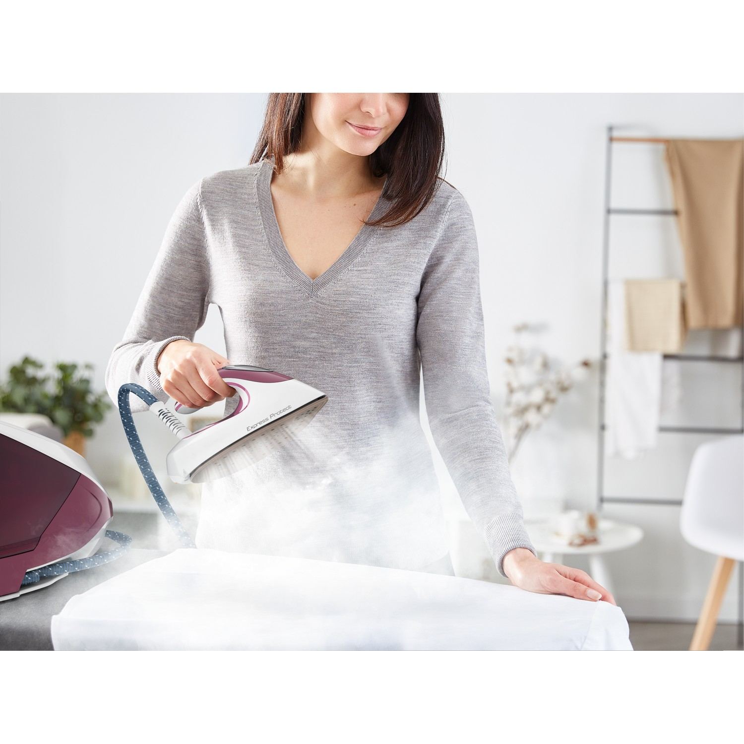 A woman irons a white shirt with a steam iron. The room has a white wall and a plant.