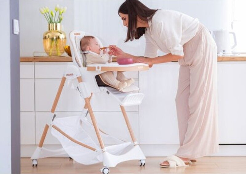 Kobieta karmi dziecko w krzesełku do karmienia. Kuchnia ma złoty wazon i białe szafki.