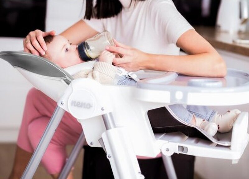 Dziecko w krzesełku do karmienia jest karmione butelką przez kobietę. Białe i szare krzesło, jasna kuchnia.