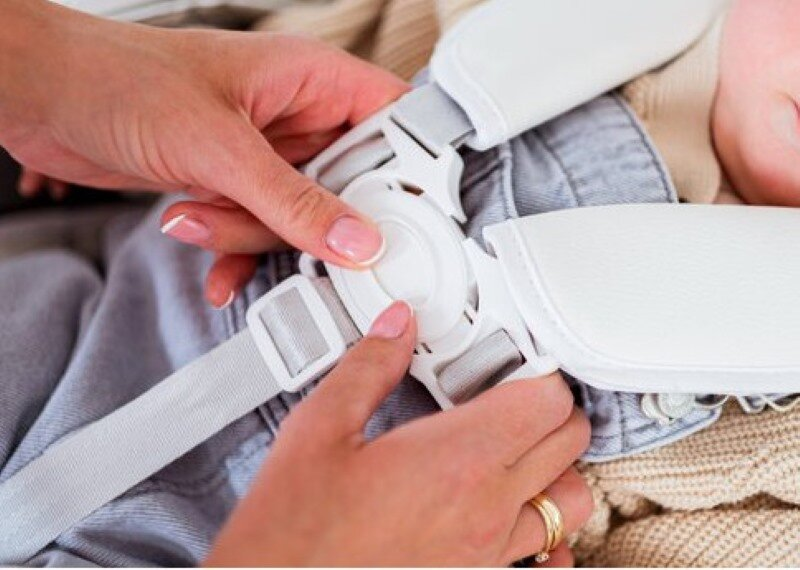 Ręce osoby zabezpieczają uprząż na klatkę piersiową dziecka. Uprząż jest biała.