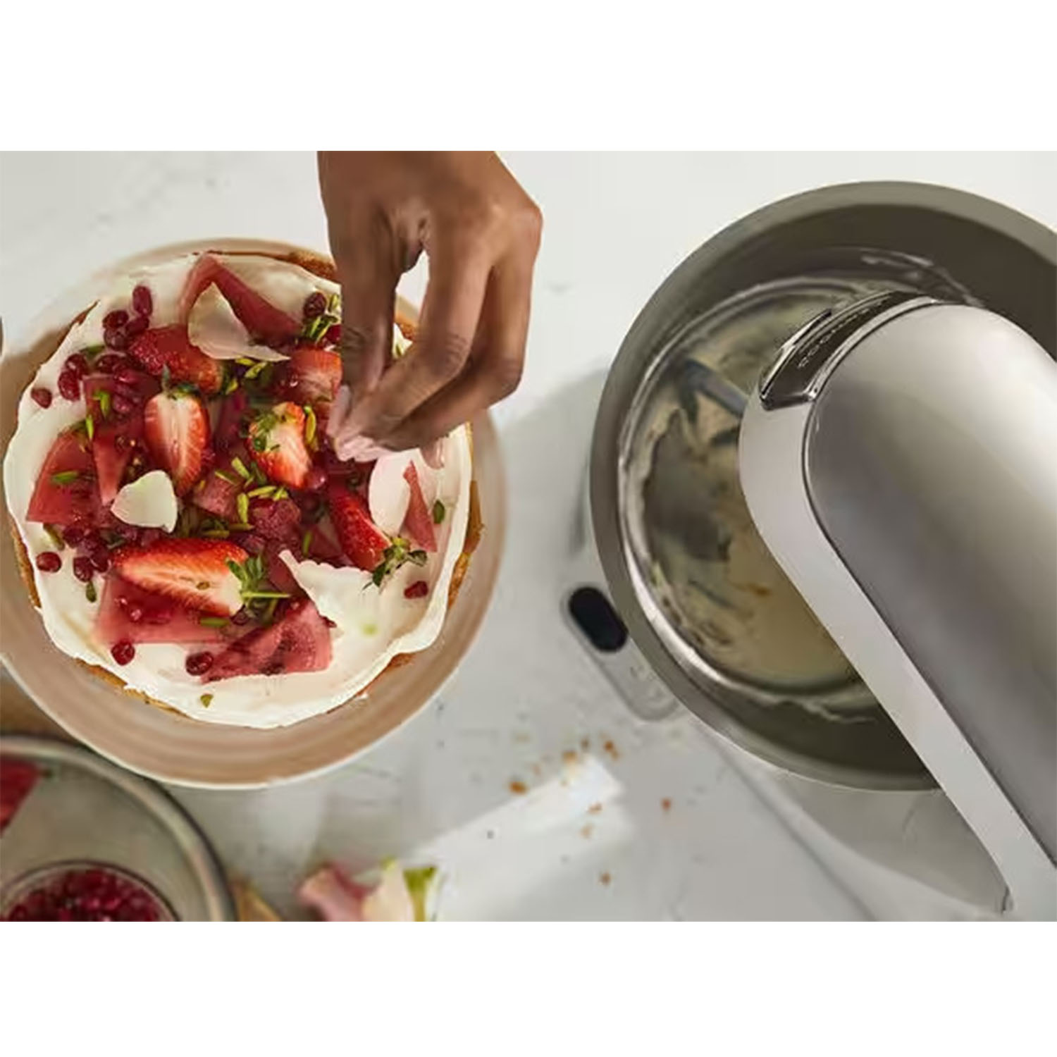 Una mano decora una torta con fragole e altri condimenti accanto a un mixer.
