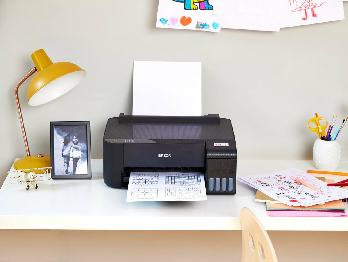 Czarna drukarka Epson na biurku z wydrukowanymi papierami i materiałami eksploatacyjnymi.