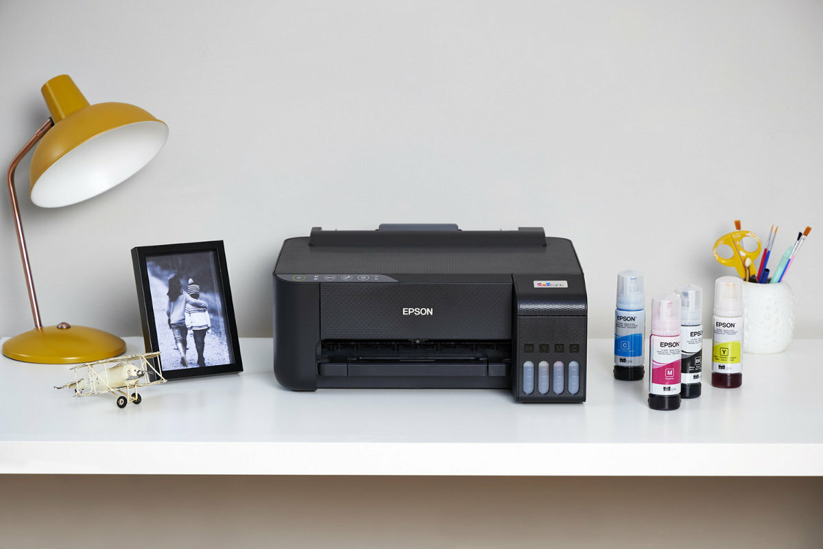 Drukarka Epson na białym biurku z wkładami atramentowymi, lampą i oprawionym zdjęciem.