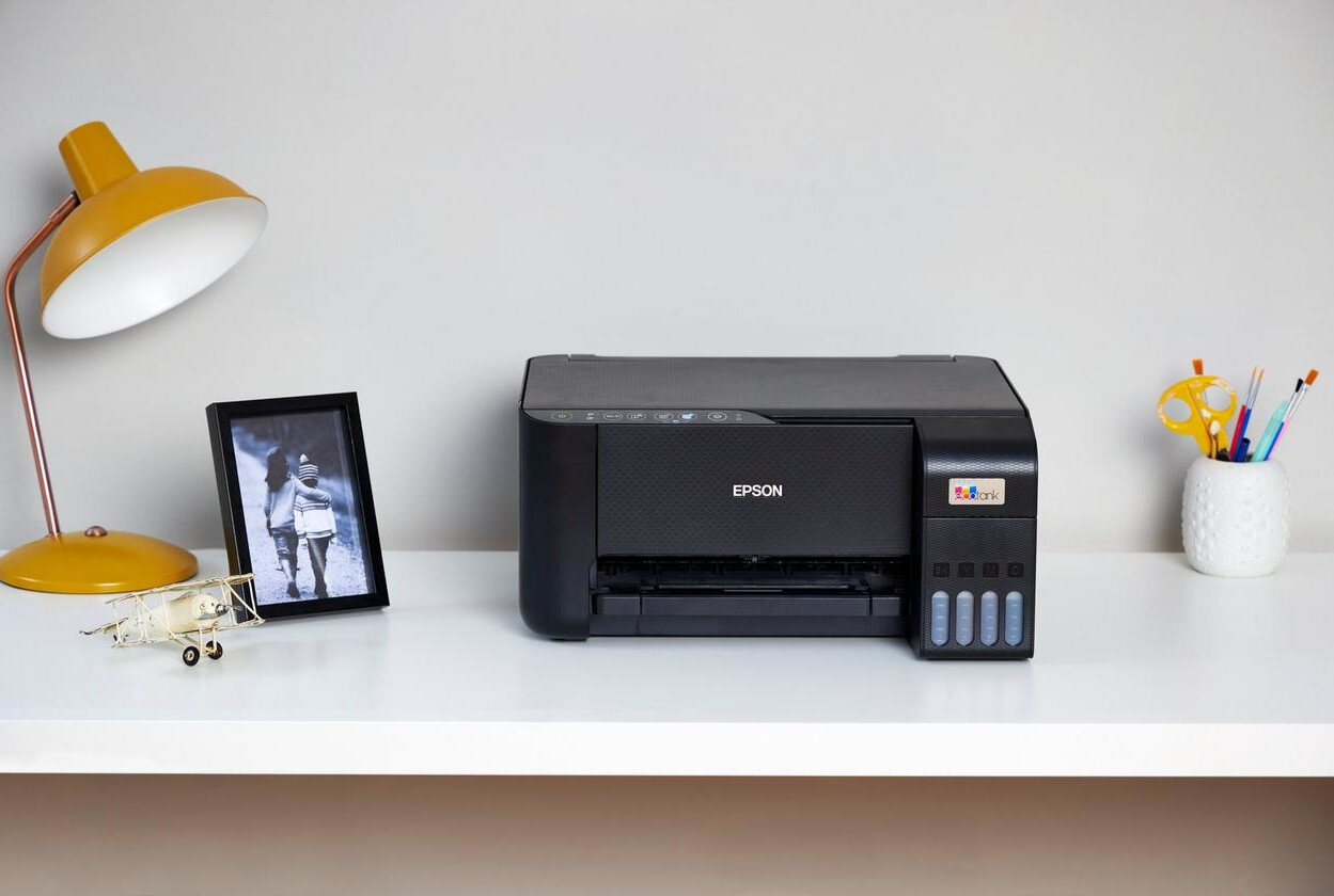 Czarna drukarka Epson na białym biurku. Lampa, ramka i ołówki obok.