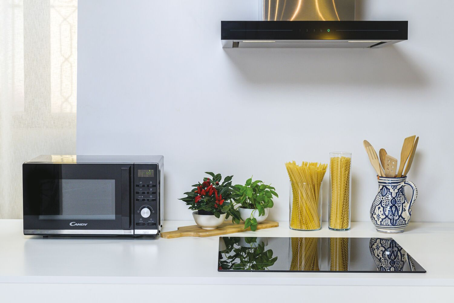 Una encimera de cocina con microondas, plantas, pasta y vitrocerámica. Fondo blanco.