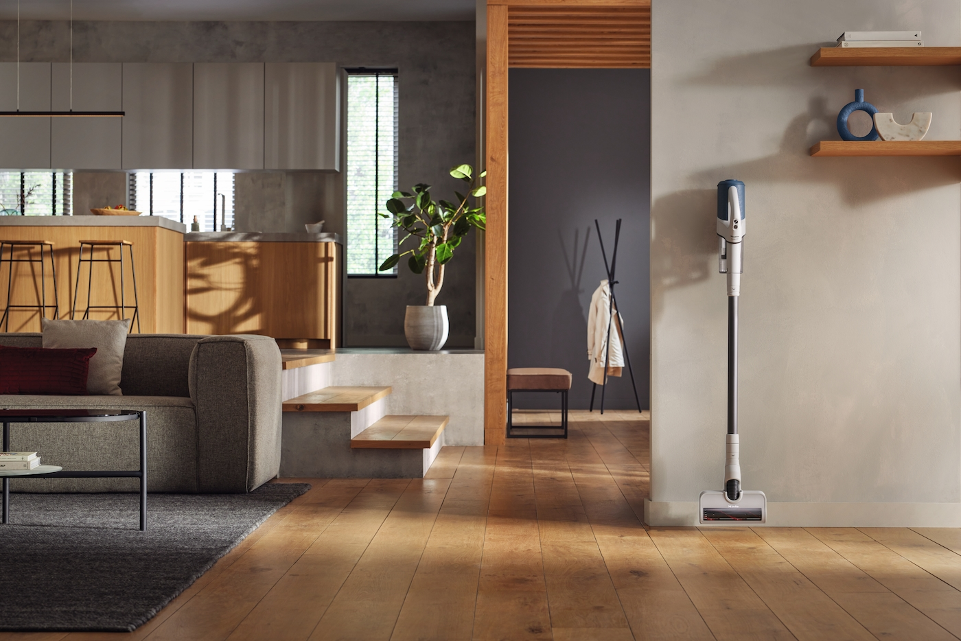 Modernes Wohnzimmer mit Sofa, Kücheninsel, Pflanze, Garderobe und Staubsauger an der Wand.