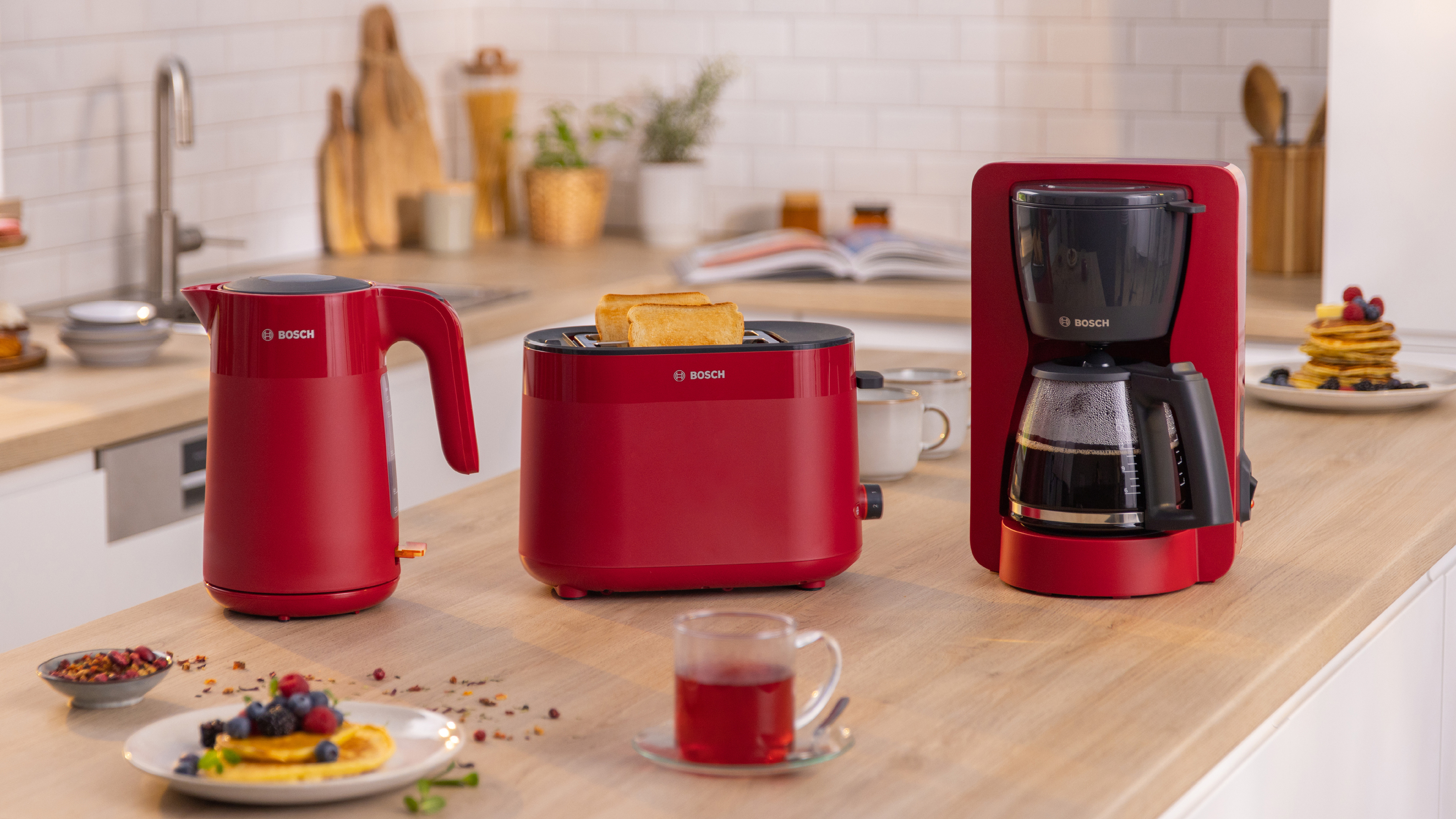Ein roter Bosch-Wasserkocher, Toaster und Kaffeemaschine auf einer Holztheke mit Lebensmitteln.