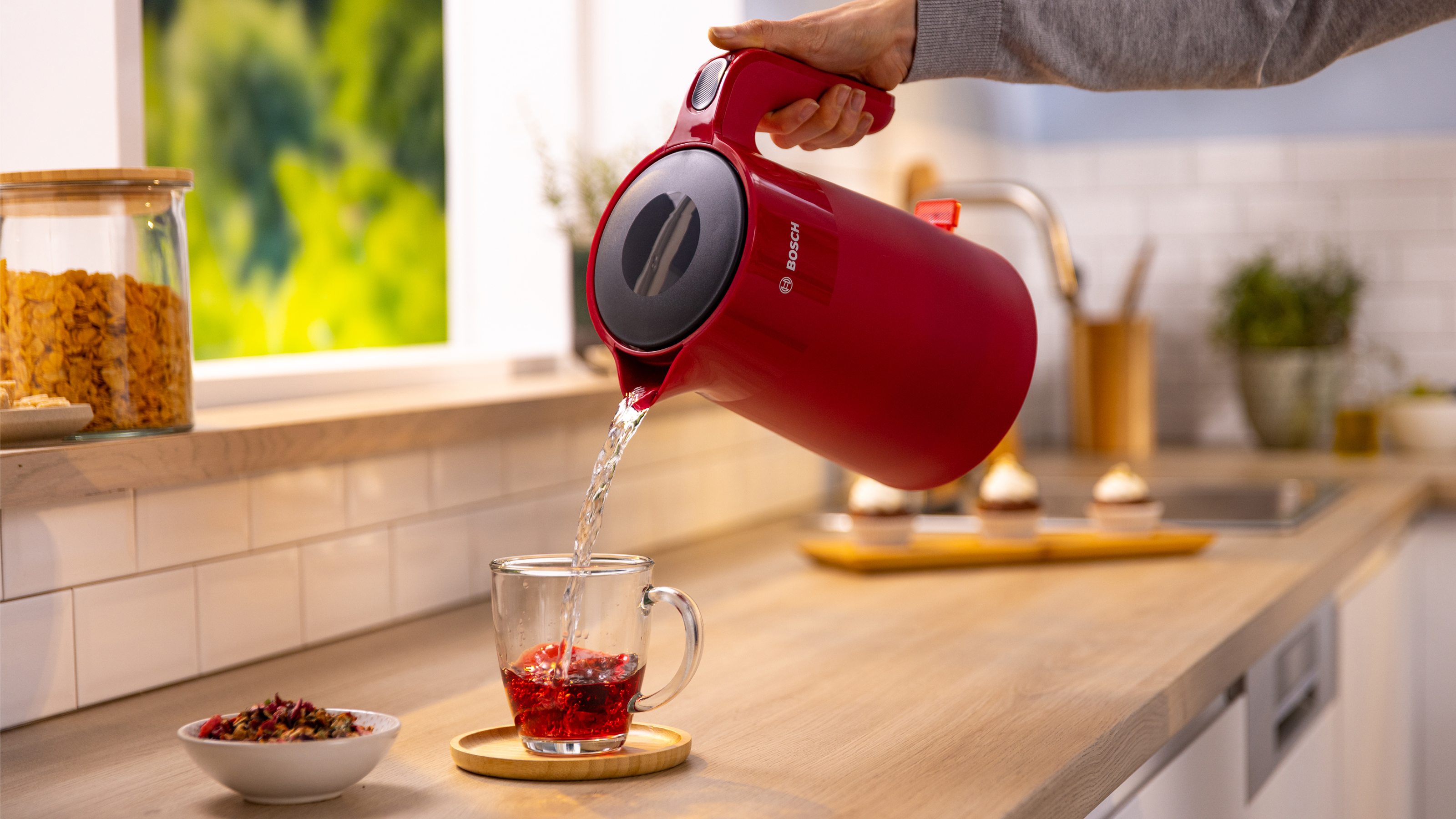 Ein roter Wasserkocher gießt Wasser in ein Glas mit Flüssigkeit. Ein Holztablett ist darunter.