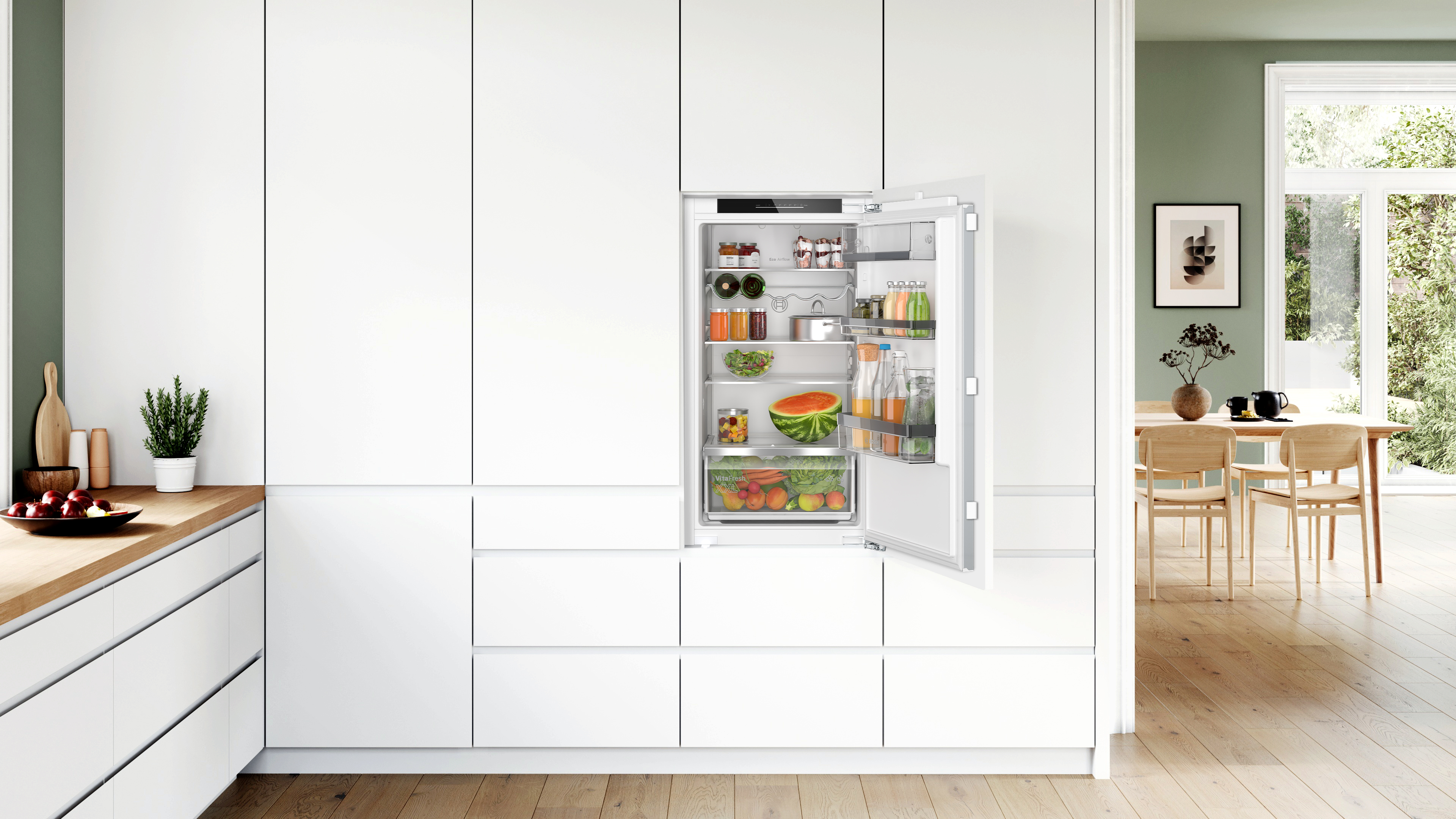 Weiße Küche mit geöffnetem Kühlschrank; Früchte, Gemüse und Lebensmittel sichtbar. Grüne Wand und Holztheke links.