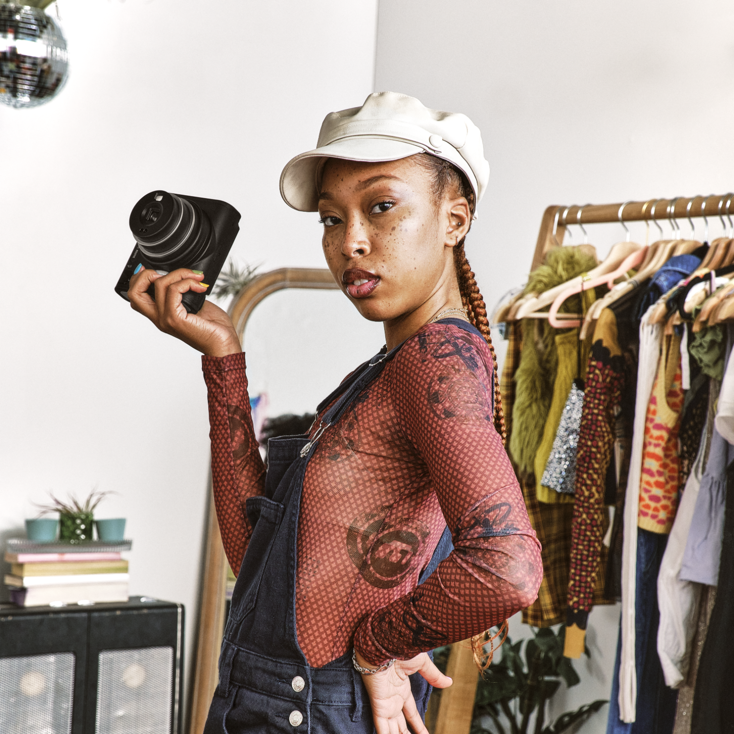 Young woman in overalls and a cap holding a camera, in a vintage clothing store.