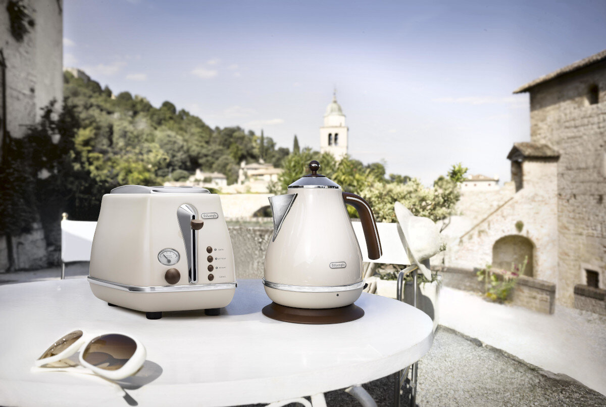 Toster i czajnik na białym stole na zewnątrz, z budynkami w tle.