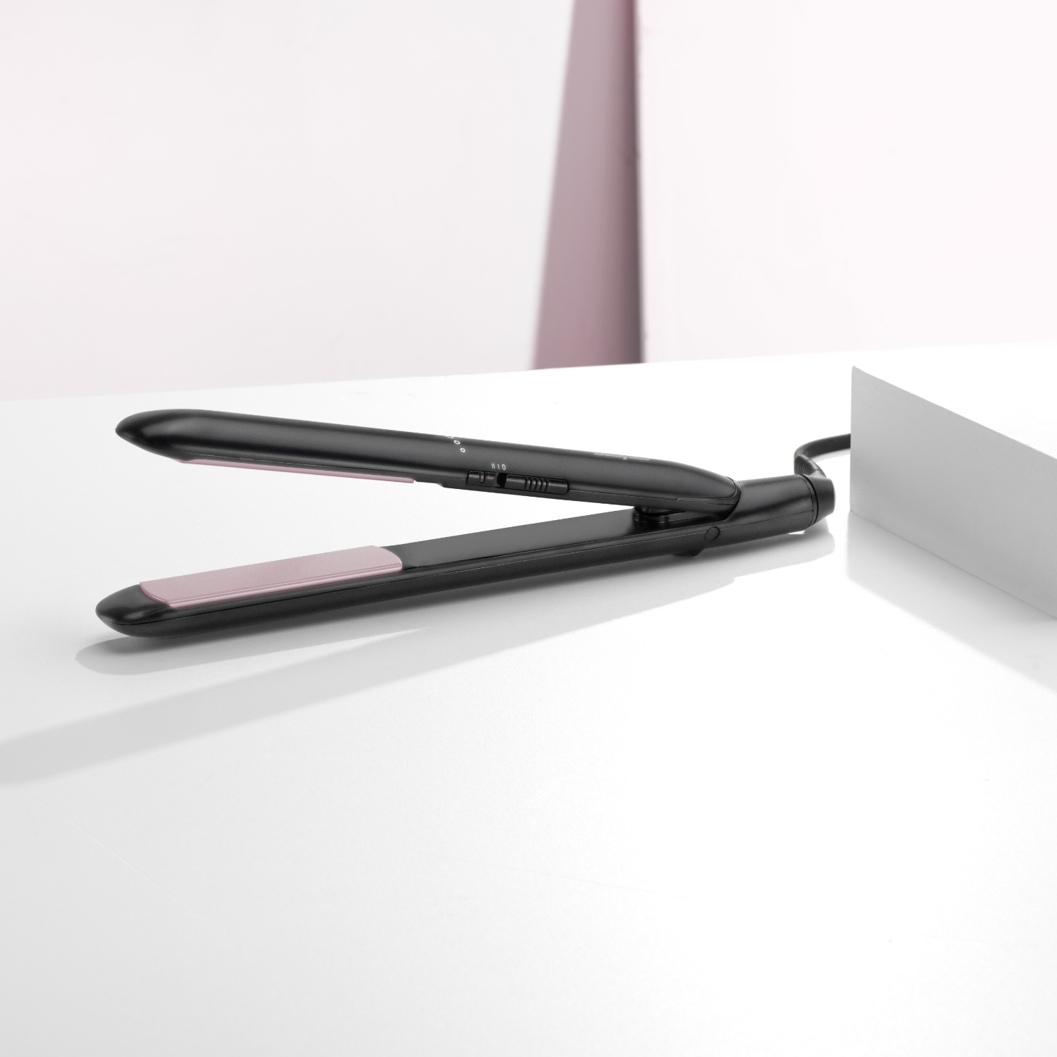 Black hair straightener on a white surface, with pink plates and a cable.