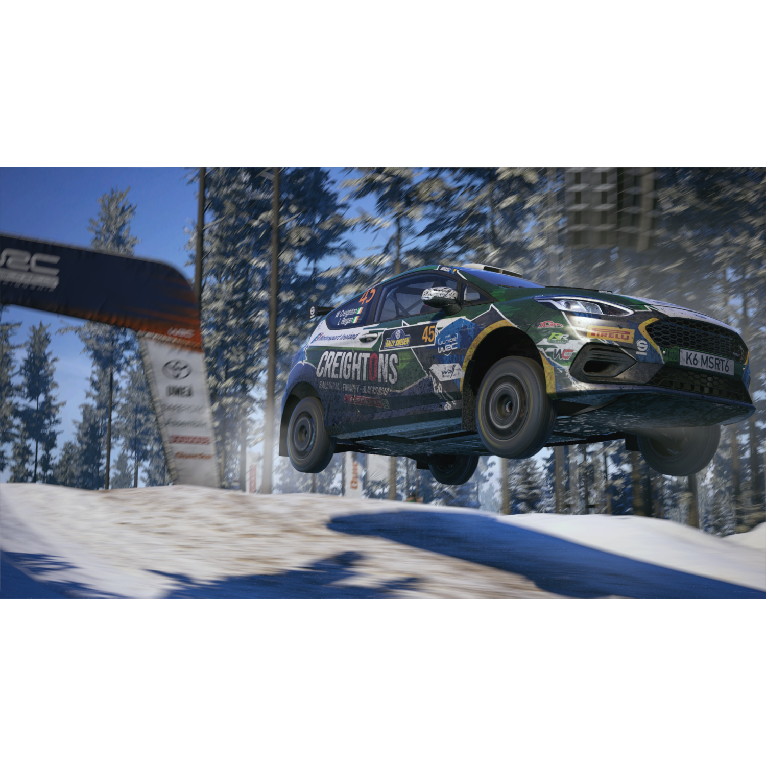 Een rallyauto die over een besneeuwde heuvel springt, met bomen aan de zijkant en een finishvlag.