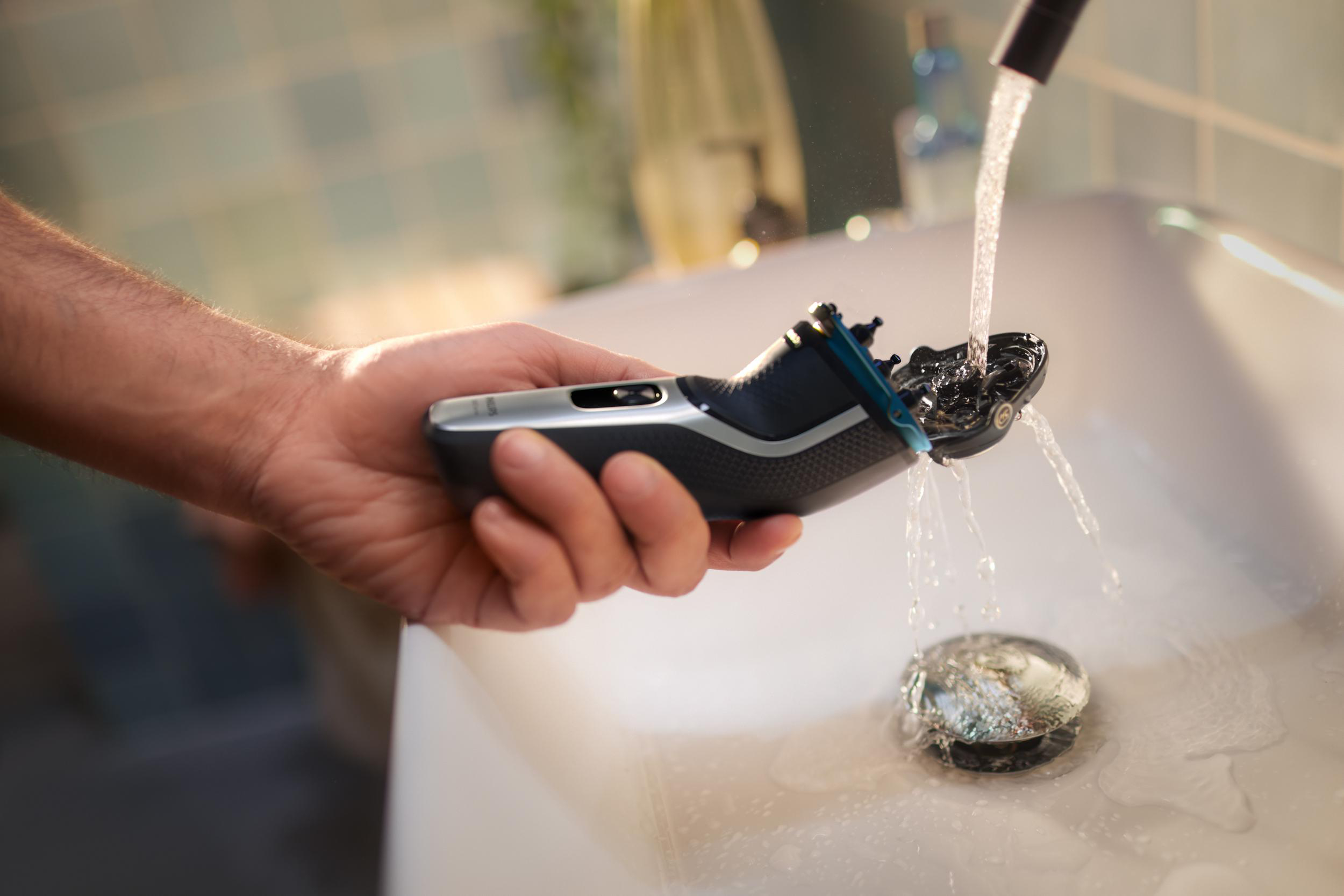 Eine Hand, die einen Rasierer unter fließendem Wasser in einem weißen Waschbecken im Badezimmer hält.