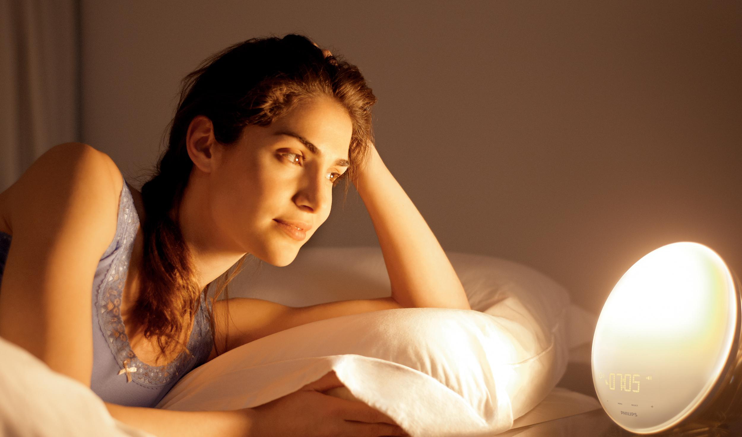 Frau im Bett blickt auf einen runden Wecker, der Raum ist schwach beleuchtet.