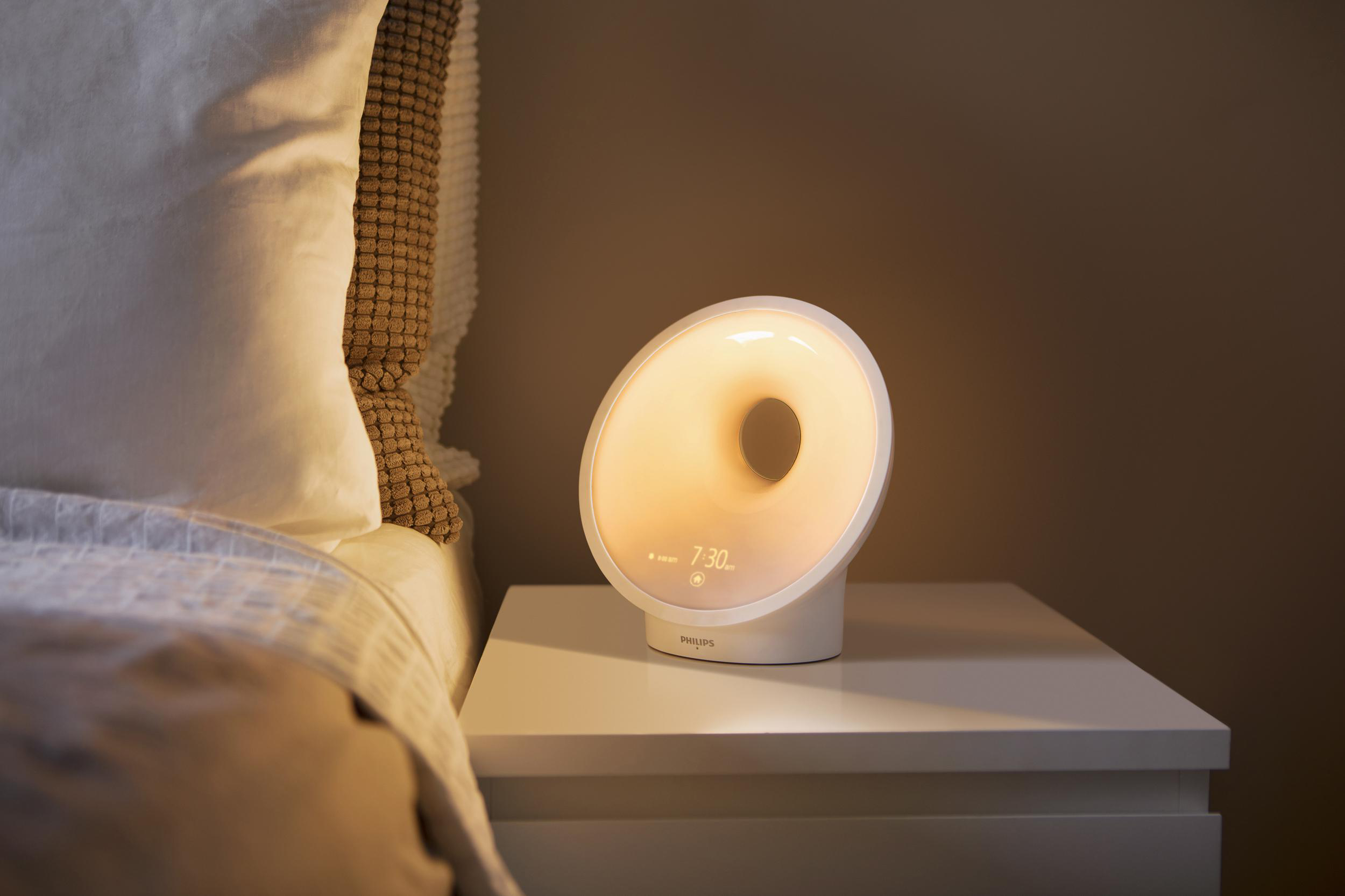 Weißes Philips Wake-Up Light auf einem Nachttisch. Es hat ein kreisförmiges Licht und eine Digitaluhr.