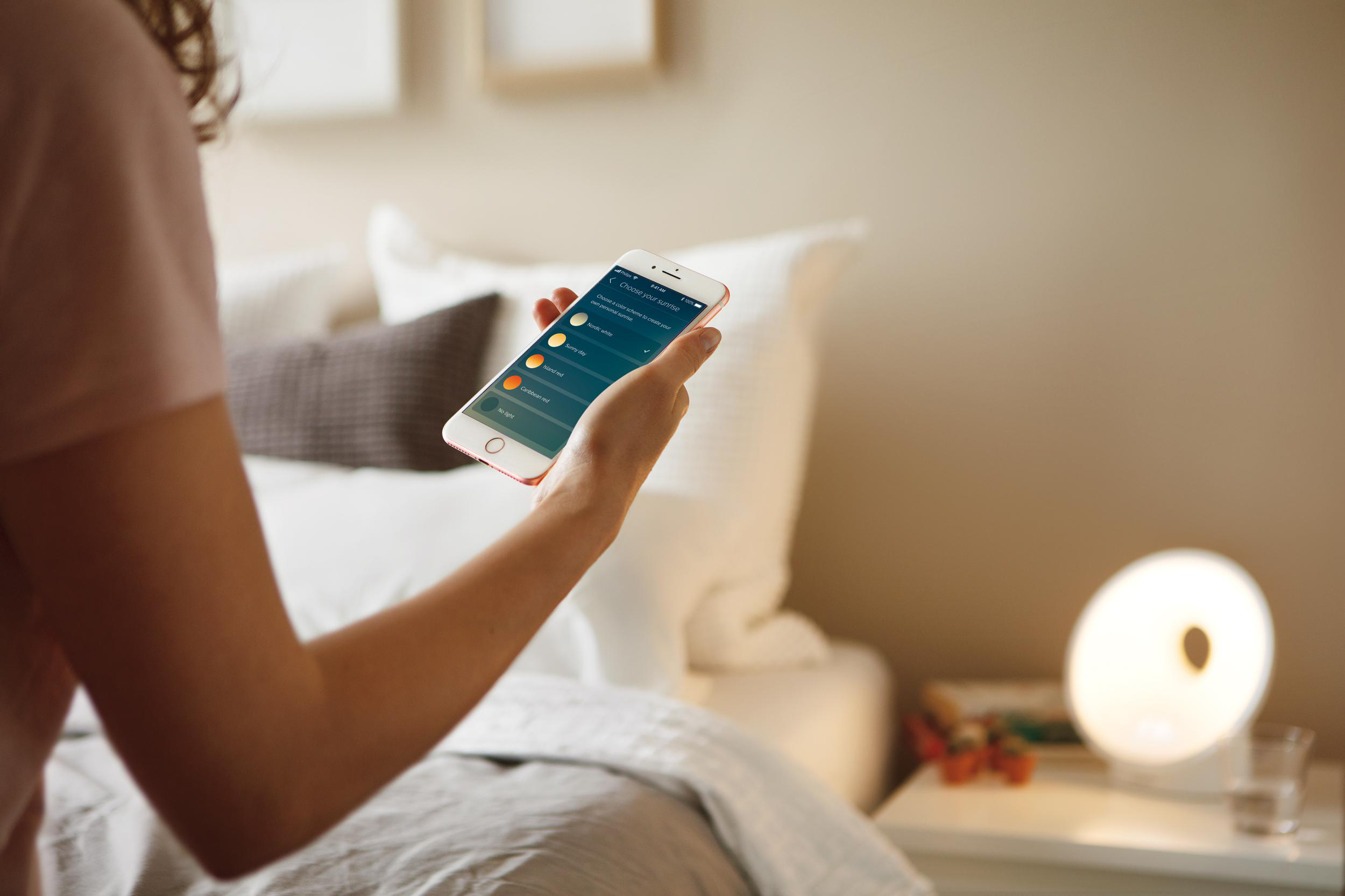 Eine Person hält ein Smartphone mit einer App zur Auswahl von Sonnenaufgangsfarben. Ein Lichtwecker steht auf dem Nachttisch.