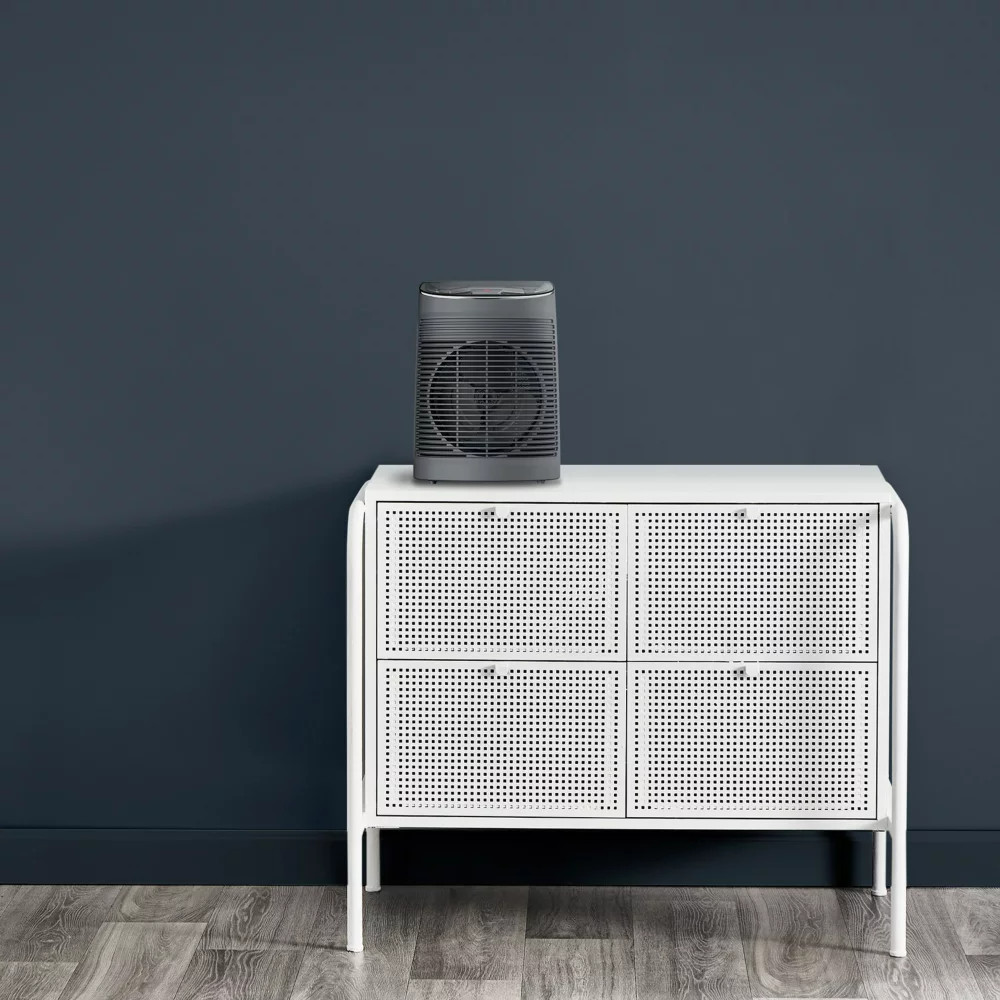 A gray fan is on a white cabinet. The cabinet has four drawers. The background is a dark blue wall.