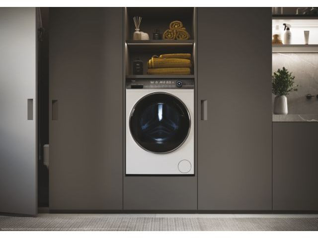 Een witte wasmachine is ingebouwd in een donkergrijze kast. Handdoeken en decoraties erboven.