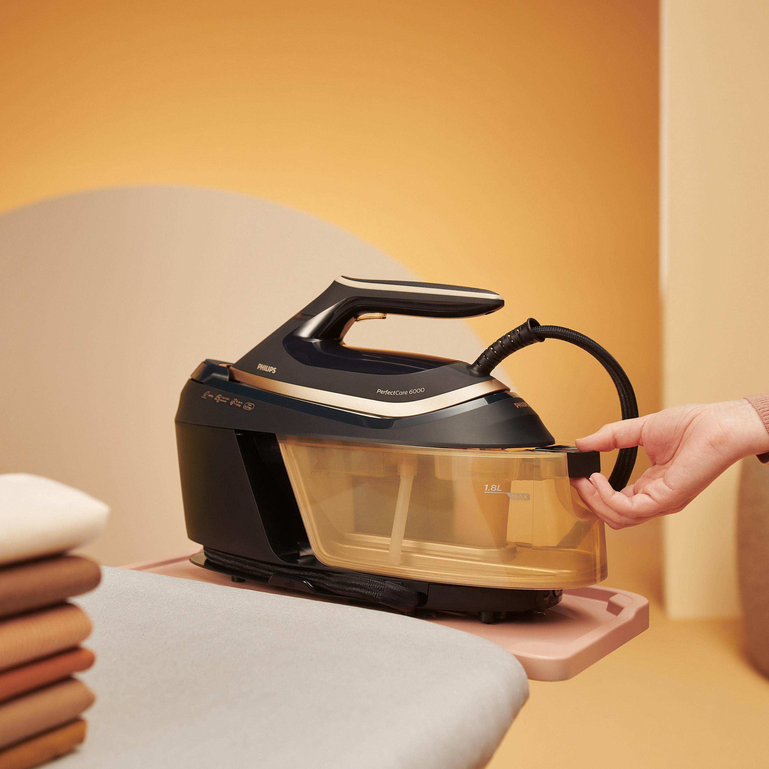 Bügelmaschine auf rosa Tablett, Hand gießt Wasser in den Tank. Beige Wand und gebügelte Kleidung sichtbar.