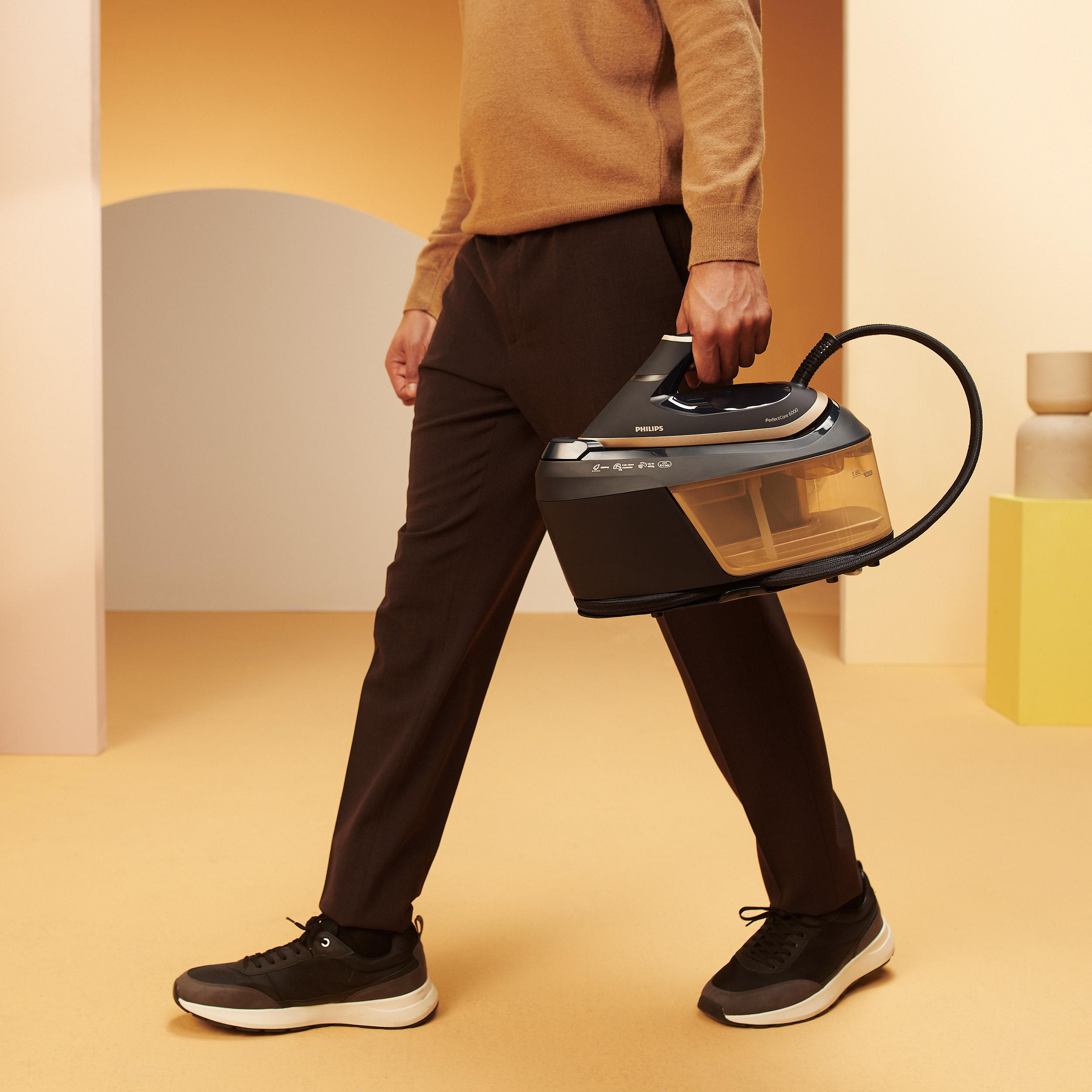 Ein Mann trägt eine Bügelmaschine, geht. Er trägt braune Hose und Schuhe. Hintergrund ist orange und pink.