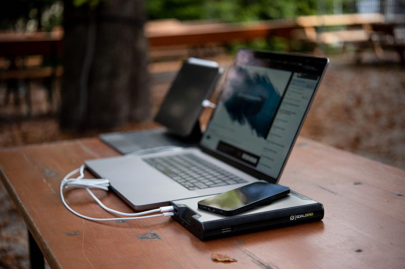 Laptop, Telefon und Powerbank auf einem Holztisch, im Freien.