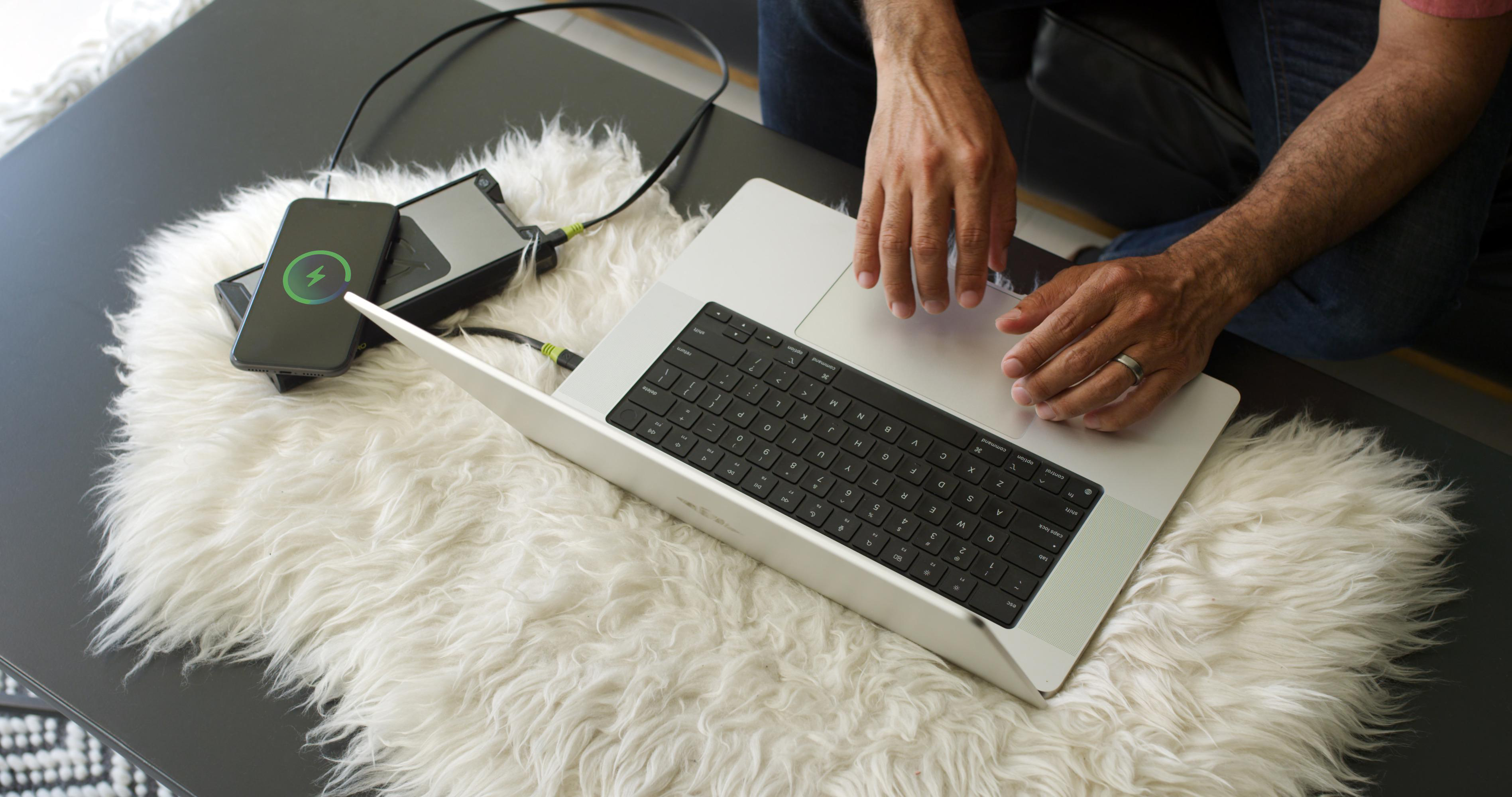 Person arbeitet an einem Laptop mit ladendem Telefon. Ein Ladegerät und Telefon auf einem weißen, flauschigen Teppich.