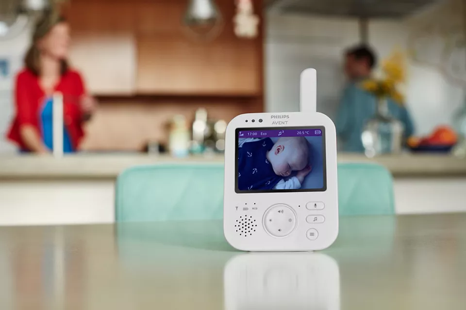 Babyphone auf einem Tisch. Ein Baby schläft im Kinderbett auf dem Bildschirm. Personen im Hintergrund.