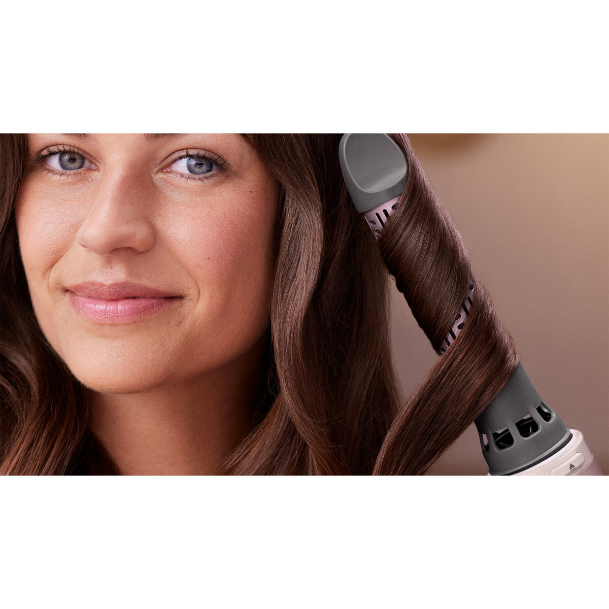 Kobieta z brązowymi włosami kręci je lokówką, uśmiechając się do kamery.