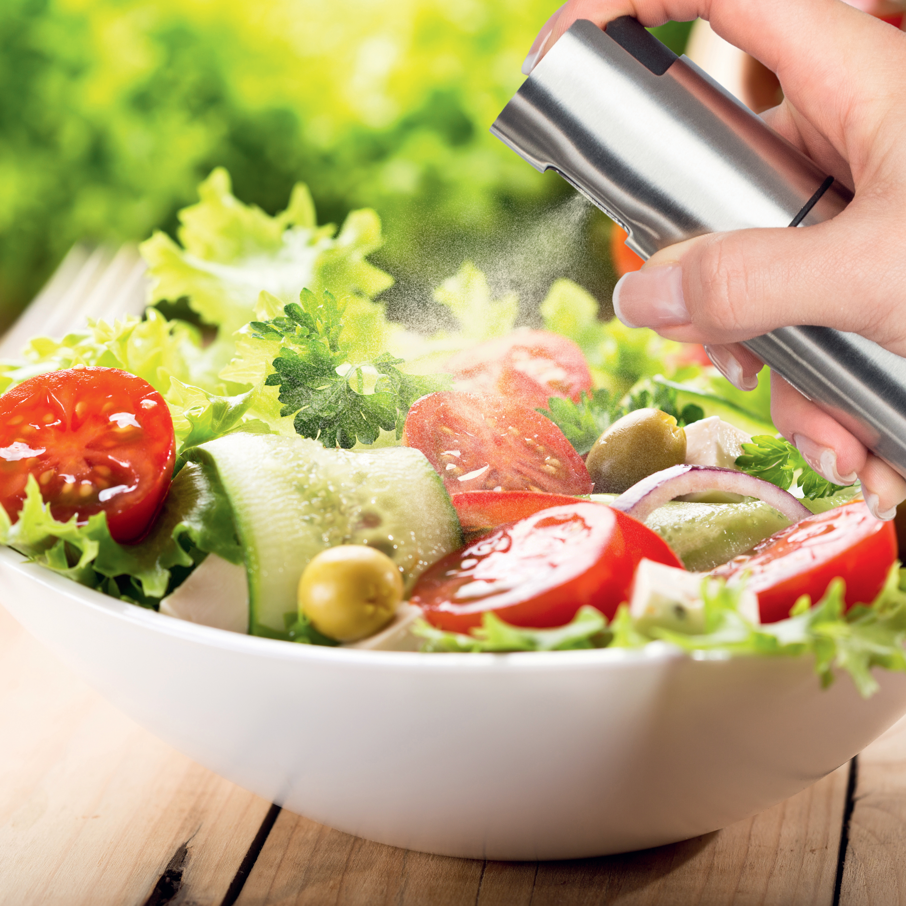 Hand sprüht Öl auf einen Salat mit Tomaten, Gurken, Oliven und Salat in einer weißen Schüssel.