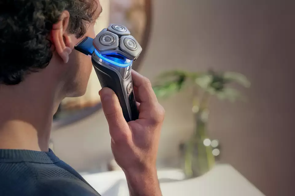Egy férfi elektromos borotvával borotválja az arcát. A borotva kék fénnyel világít és használatban van.