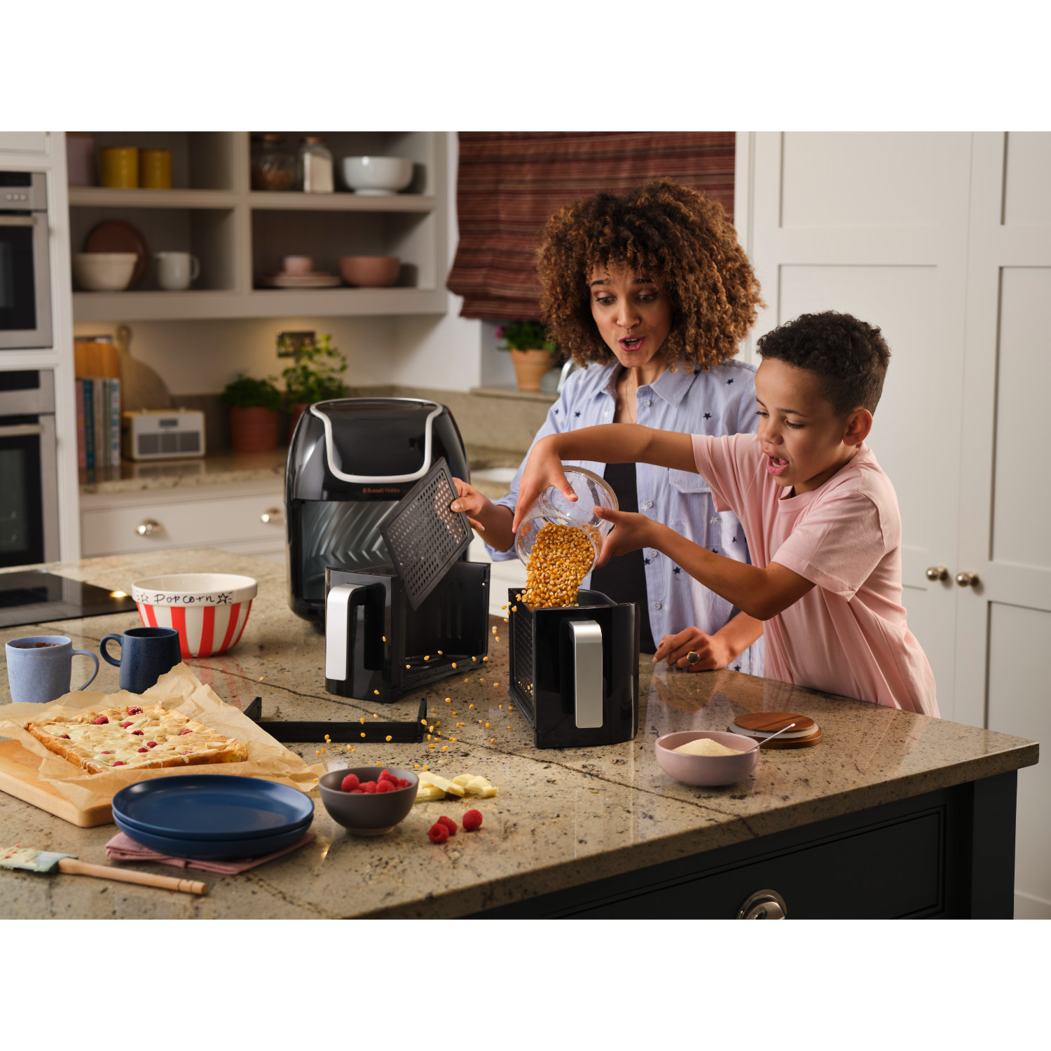 A woman and child prepare popcorn. The child pours kernels into a black machine. Other foods and kitchen items are on the counter.