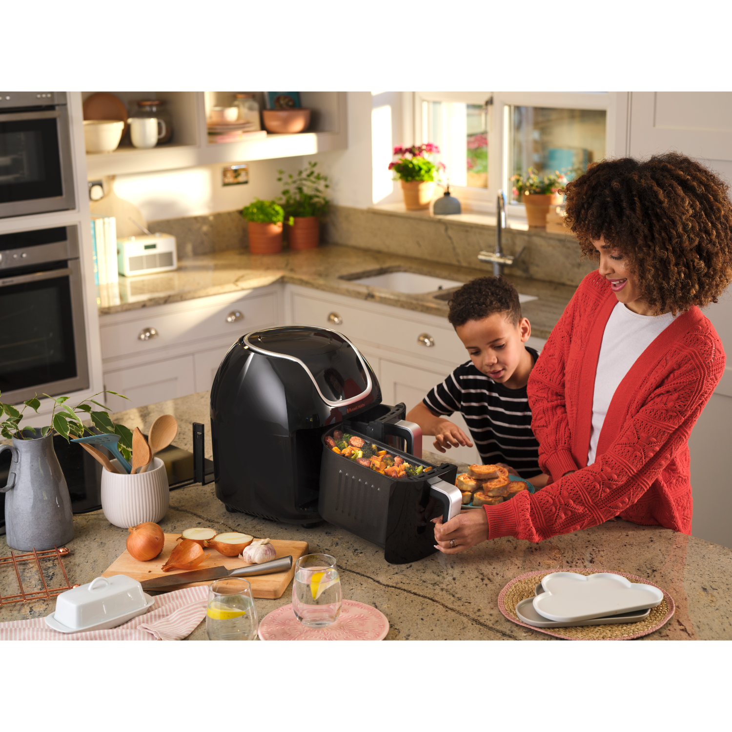 A woman and child in a kitchen with food. A black air fryer sits on the counter. Vegetables and bread are visible.