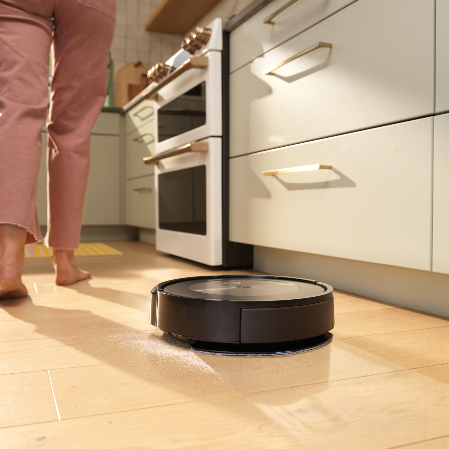 Een robotstofzuiger maakt een keuken schoon. Een persoon staat in de buurt. Kasten en een oven op de achtergrond.