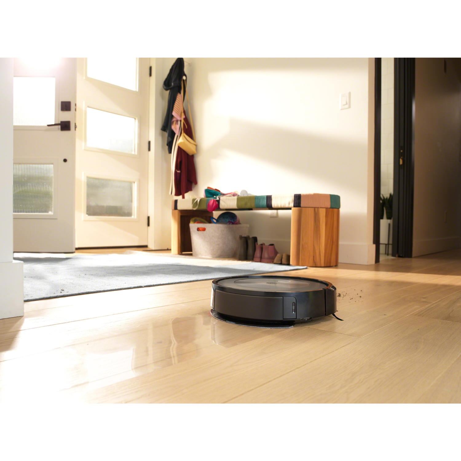 Een robotstofzuiger op een houten vloer. De scène omvat een deur, een kleed en een bank.