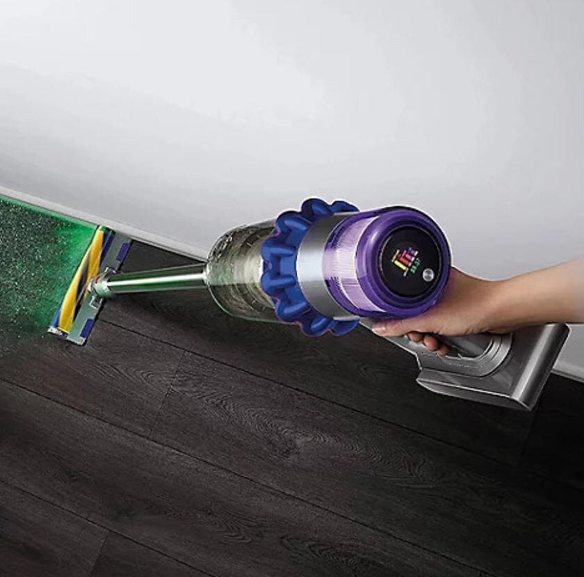 Osoba odkurza ciemną drewnianą podłogę w pobliżu ściany za pomocą bezprzewodowego odkurzacza.