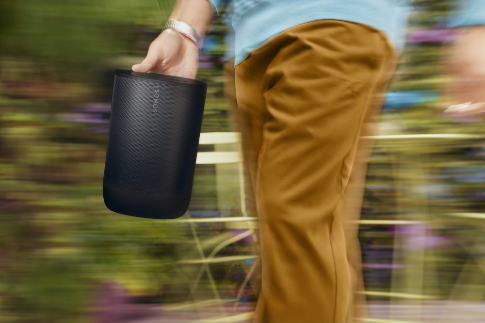 A person holding a black portable speaker walking on a blurred background of greenery.