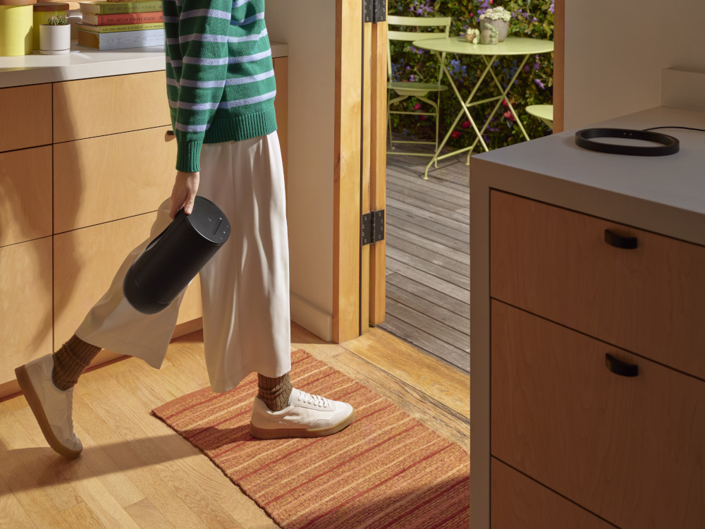 A person holding a black speaker walks towards a doorway. A red rug is on the floor. Sunny background.
