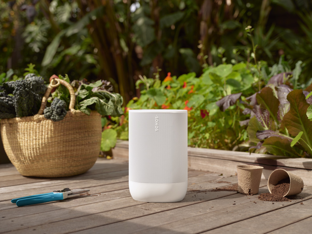 Witte speaker op houten terras met tuinplanten, mand met groen en tuingereedschap.