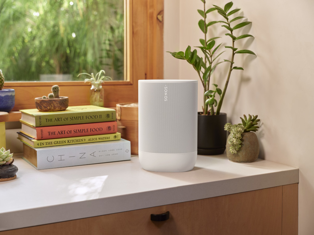 Een witte Sonos-luidspreker op een aanrecht met boeken, planten en een raam op de achtergrond.