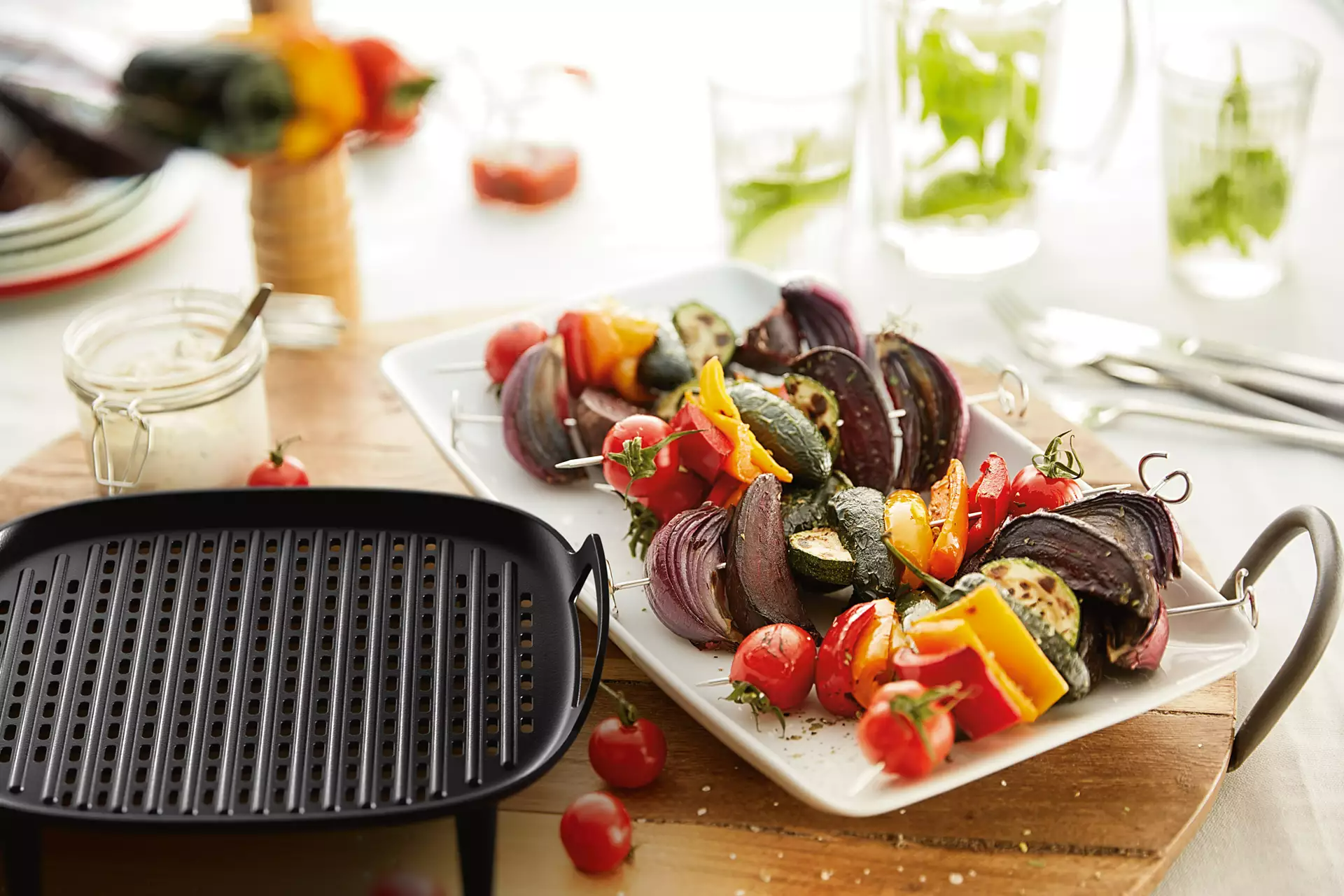 Grillezett zöldségnyársak fehér tányéron, fekete grill, szósz üveg, víz poharak a fafelületen.