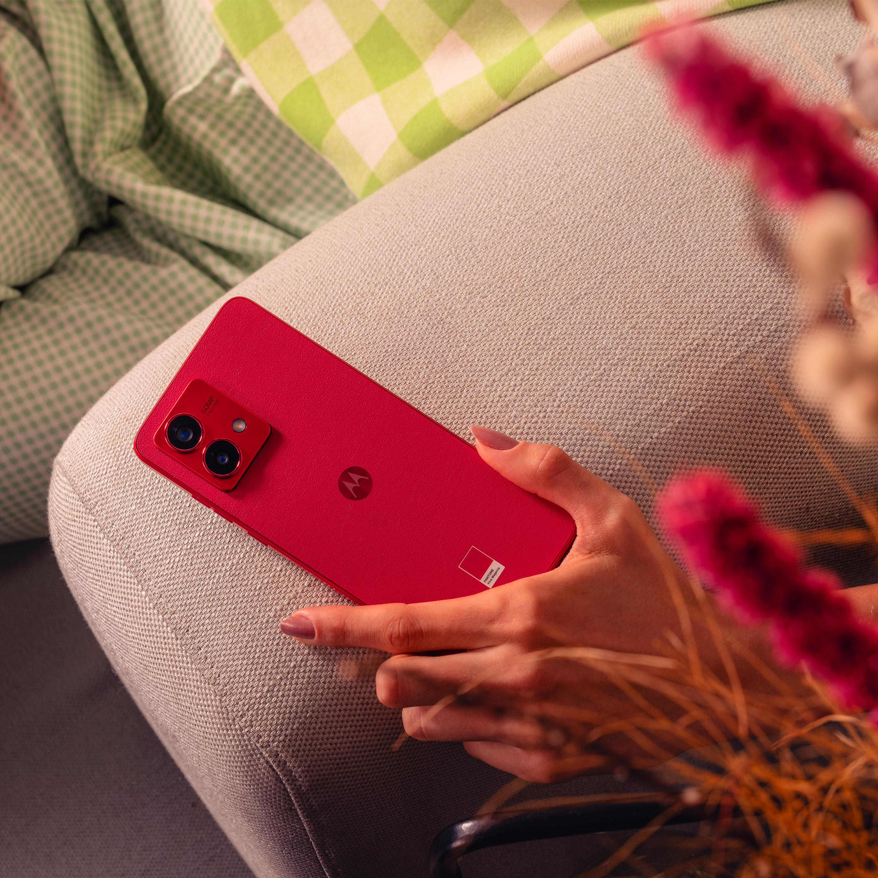 Ein rotes Motorola-Telefon wird von einer Hand gehalten, mit rosa Hintergrund und Blumen im Vordergrund.