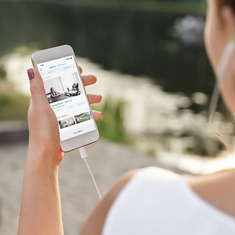 Eine Person hält ein Telefon mit einer Home-Security-App. Der Hintergrund ist eine verschwommene Ansicht eines Sees und grüner Bäume.