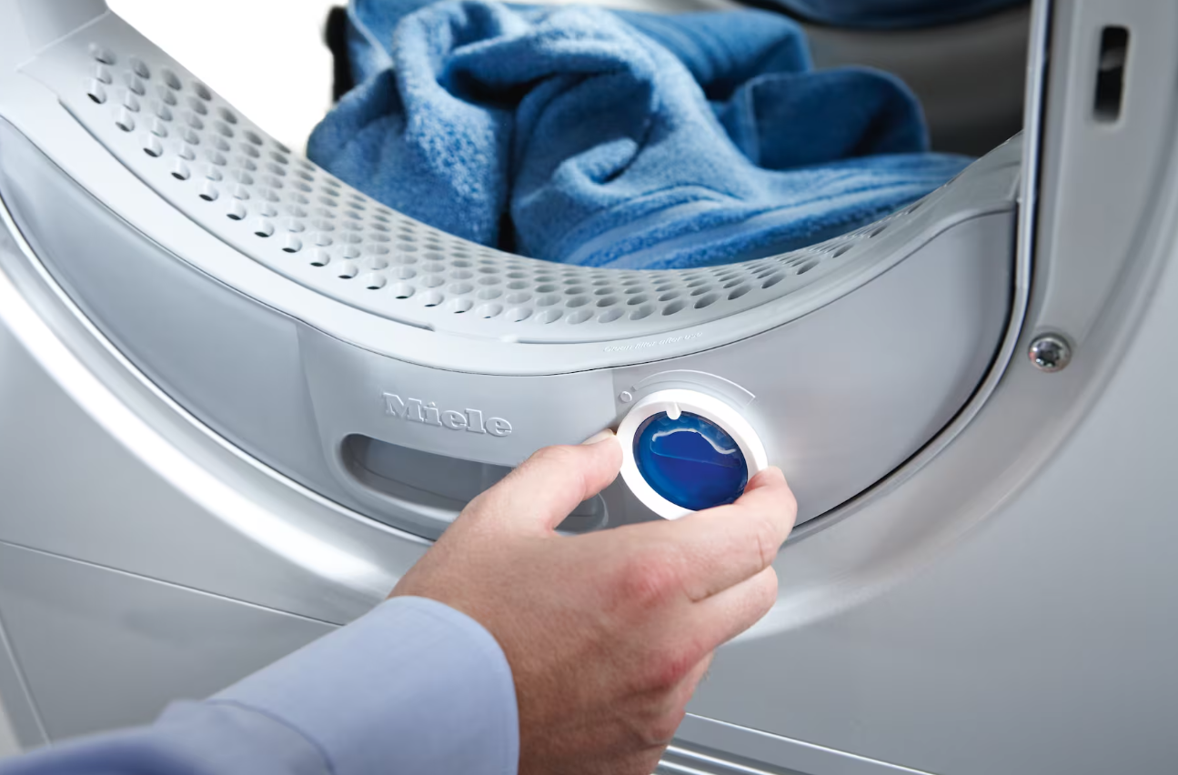 Eine Hand dreht einen Drehregler an einem Miele-Trockner. Blaue Flüssigkeit im Inneren des Reglers sichtbar.