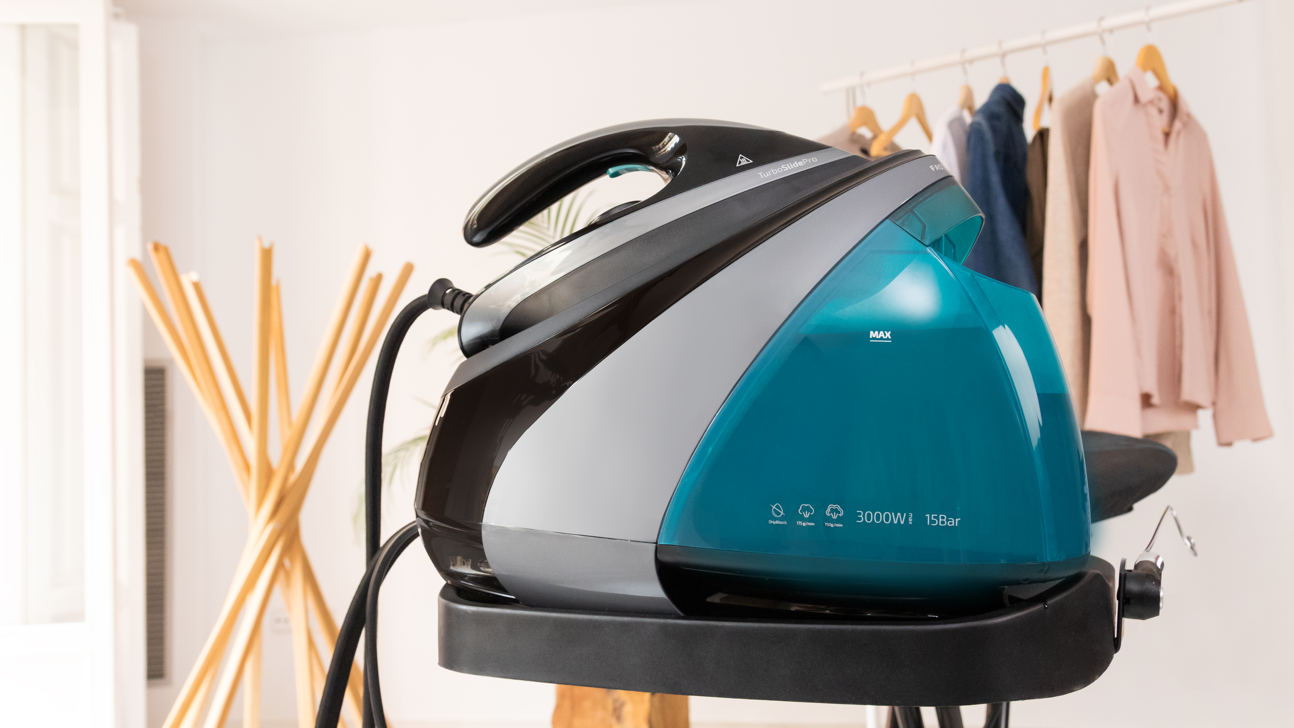 Plancha de vapor con depósito de agua, asa y cable de alimentación. Ropa en perchas al fondo.