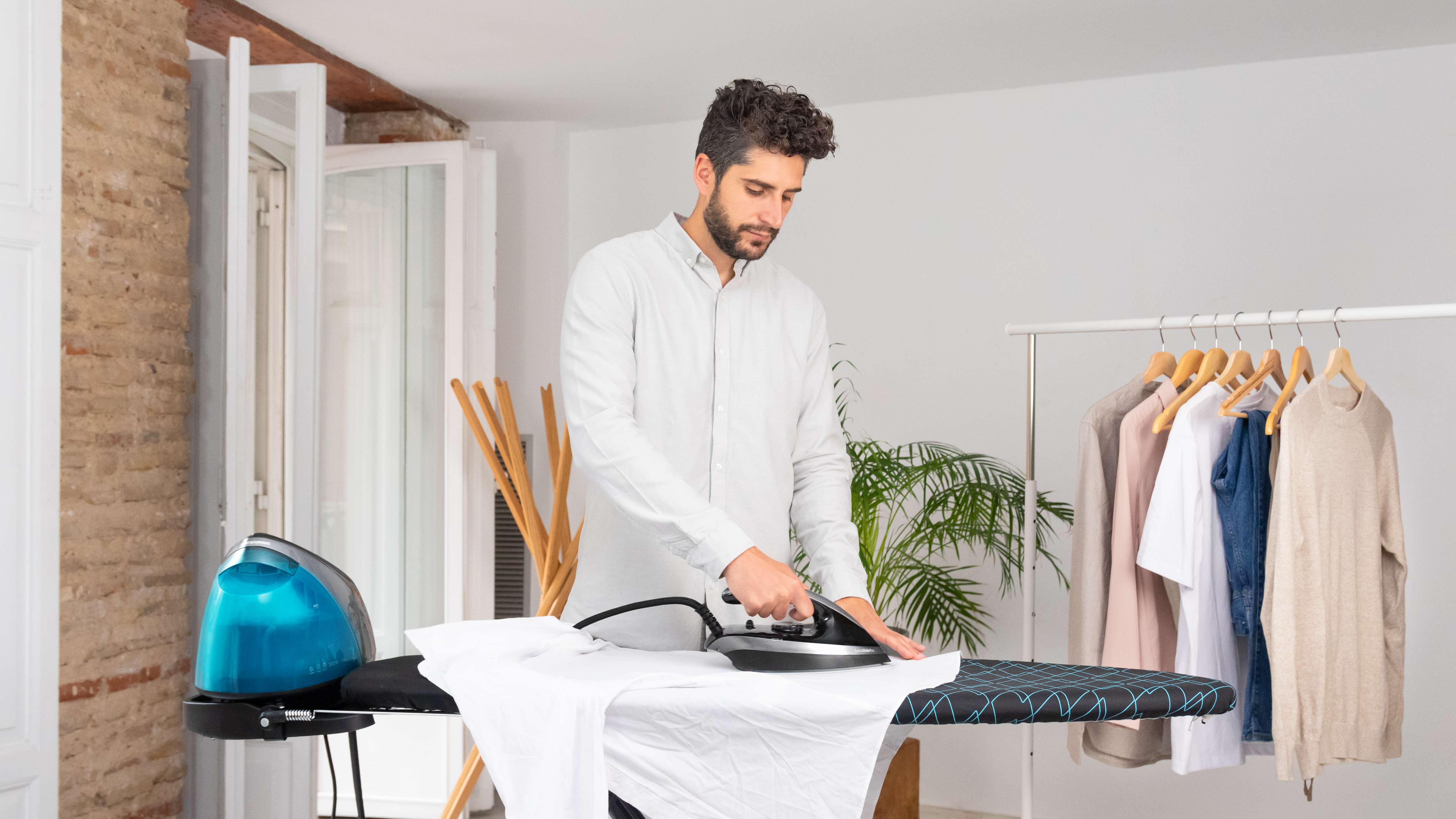 Un hombre planchando una camisa blanca sobre una tabla de planchar. Ropa colgada en un perchero al fondo.