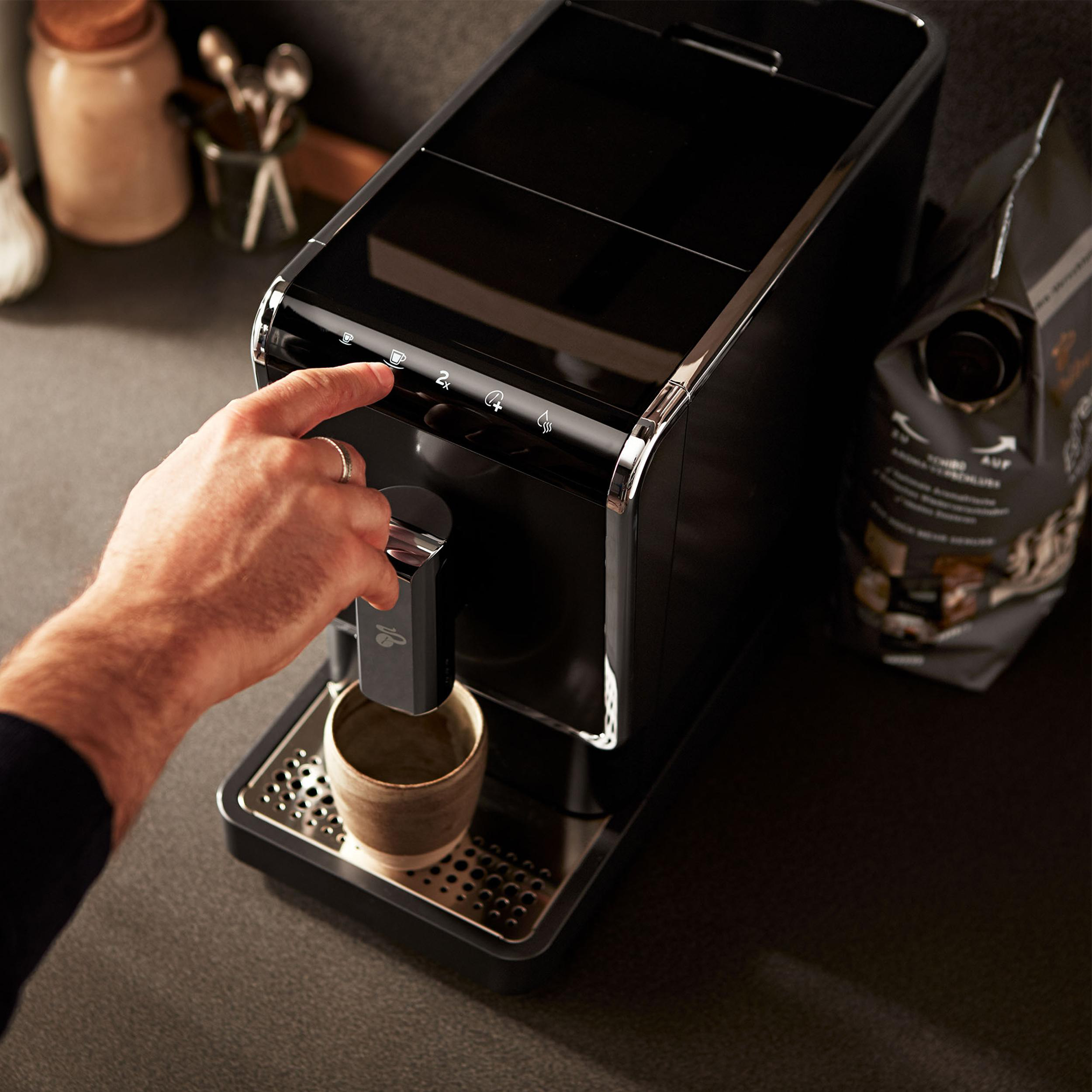 Eine schwarze Kaffeemaschine, mit einer Hand, die einen Knopf drückt, einer Tasse und einer Tüte Kaffee.